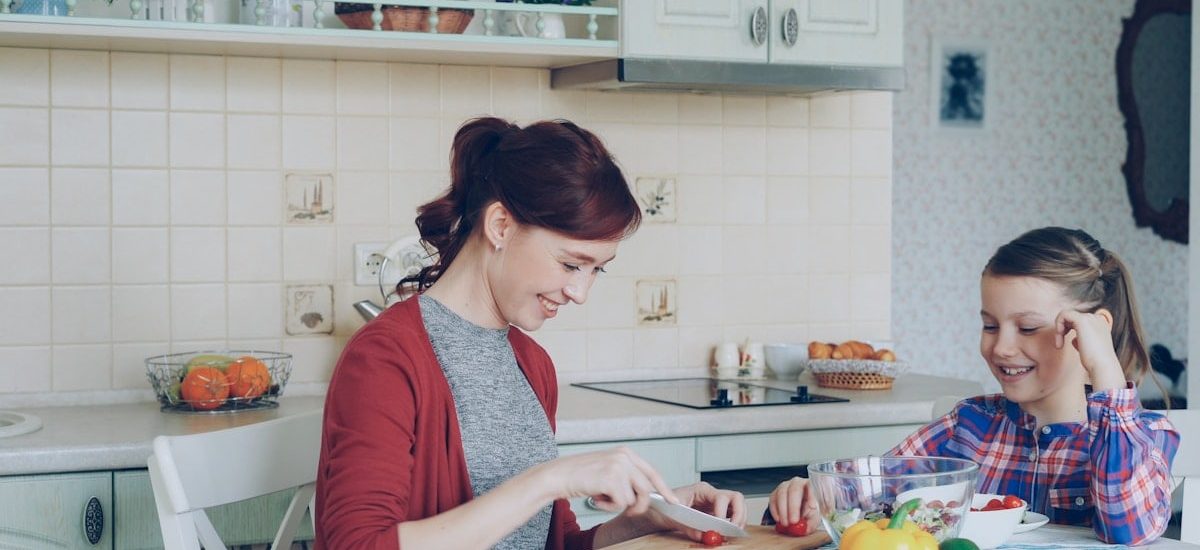 Happy young mother cooking vegetables cutting tomatoes and talking to her cheerful daughter in the kitchen at home. Family, cook, and people concept