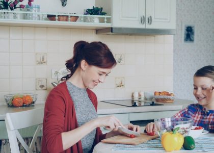 Happy young mother cooking vegetables cutting tomatoes and talking to her cheerful daughter in the kitchen at home. Family, cook, and people concept
